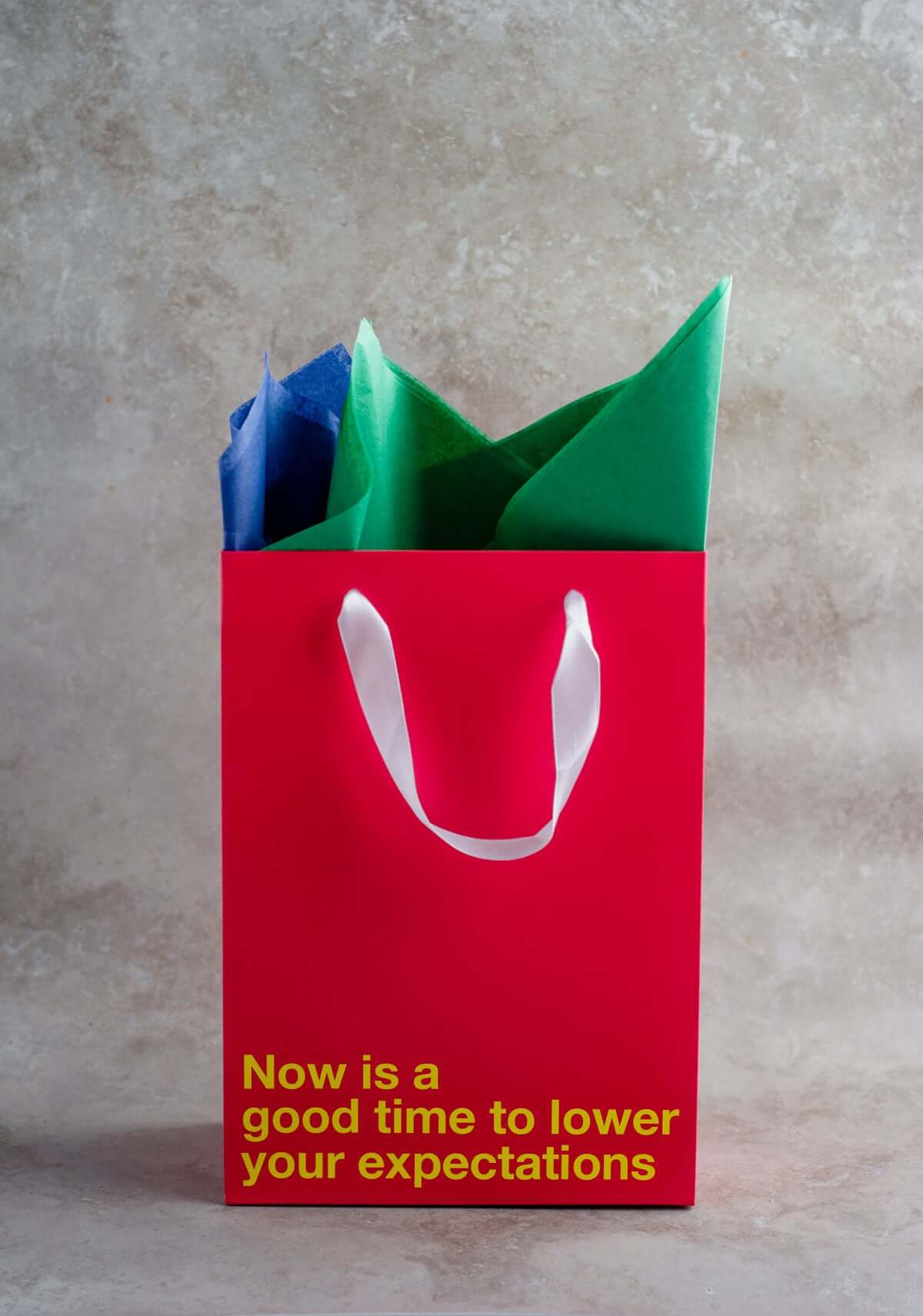 Funny gift bag in red with colorful tissue paper, featuring the text 'Now is a good time to lower your expectations'.