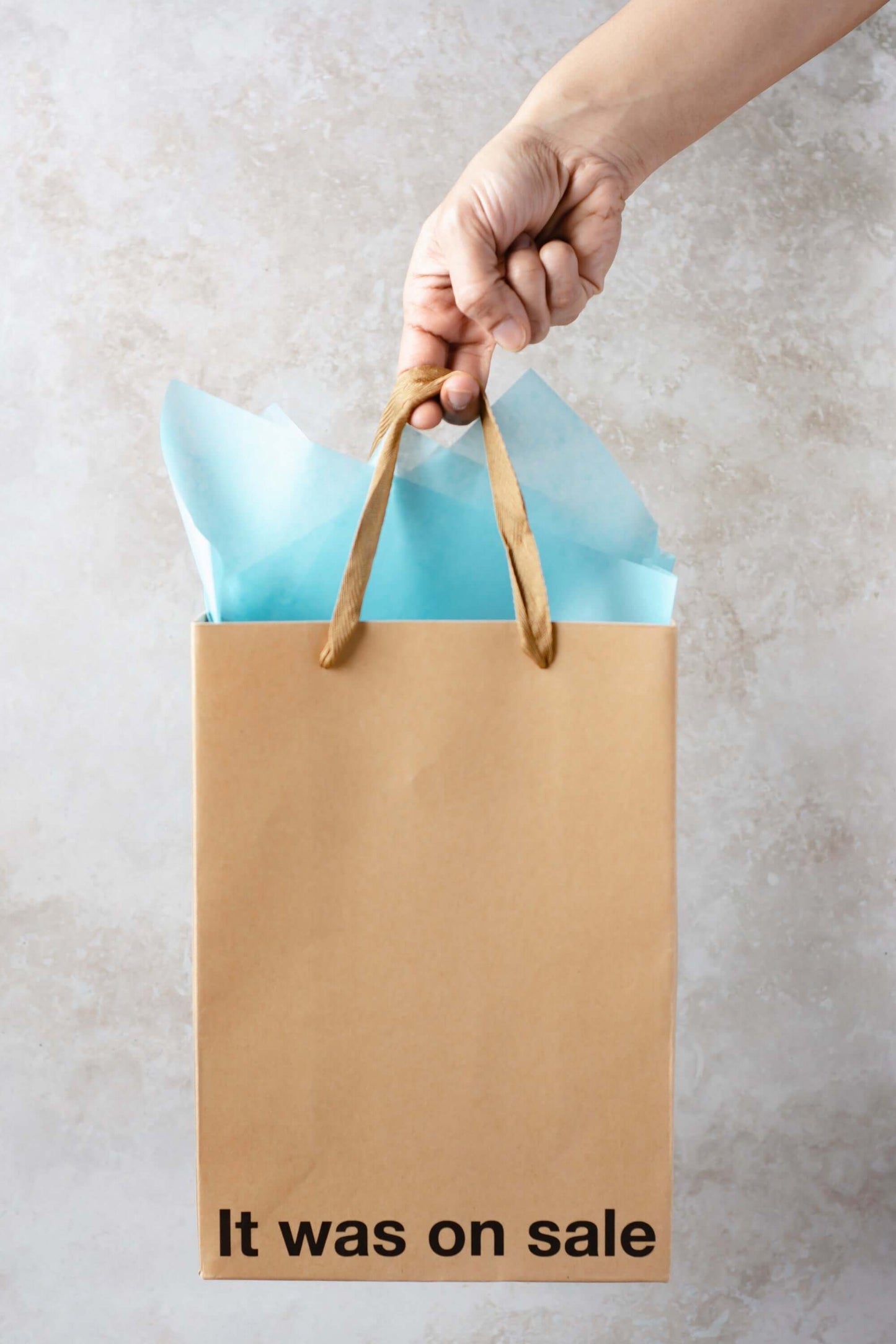 Funny gift bag held in a hand featuring a kraft design with the text 'It was on sale' and blue tissue paper.