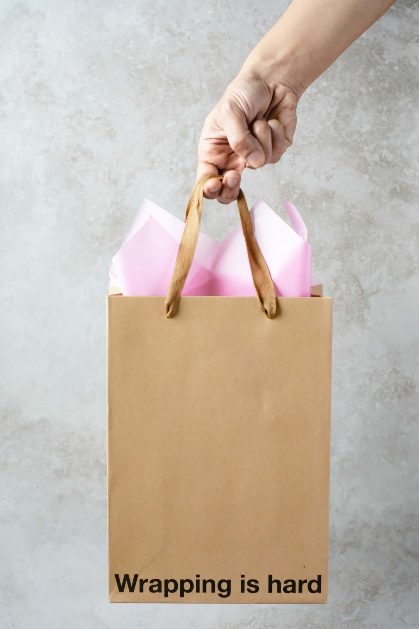 Hand holding a kraft gift bag with the text 'Wrapping is hard' and pink tissue paper inside, perfect for a funny gift bag.