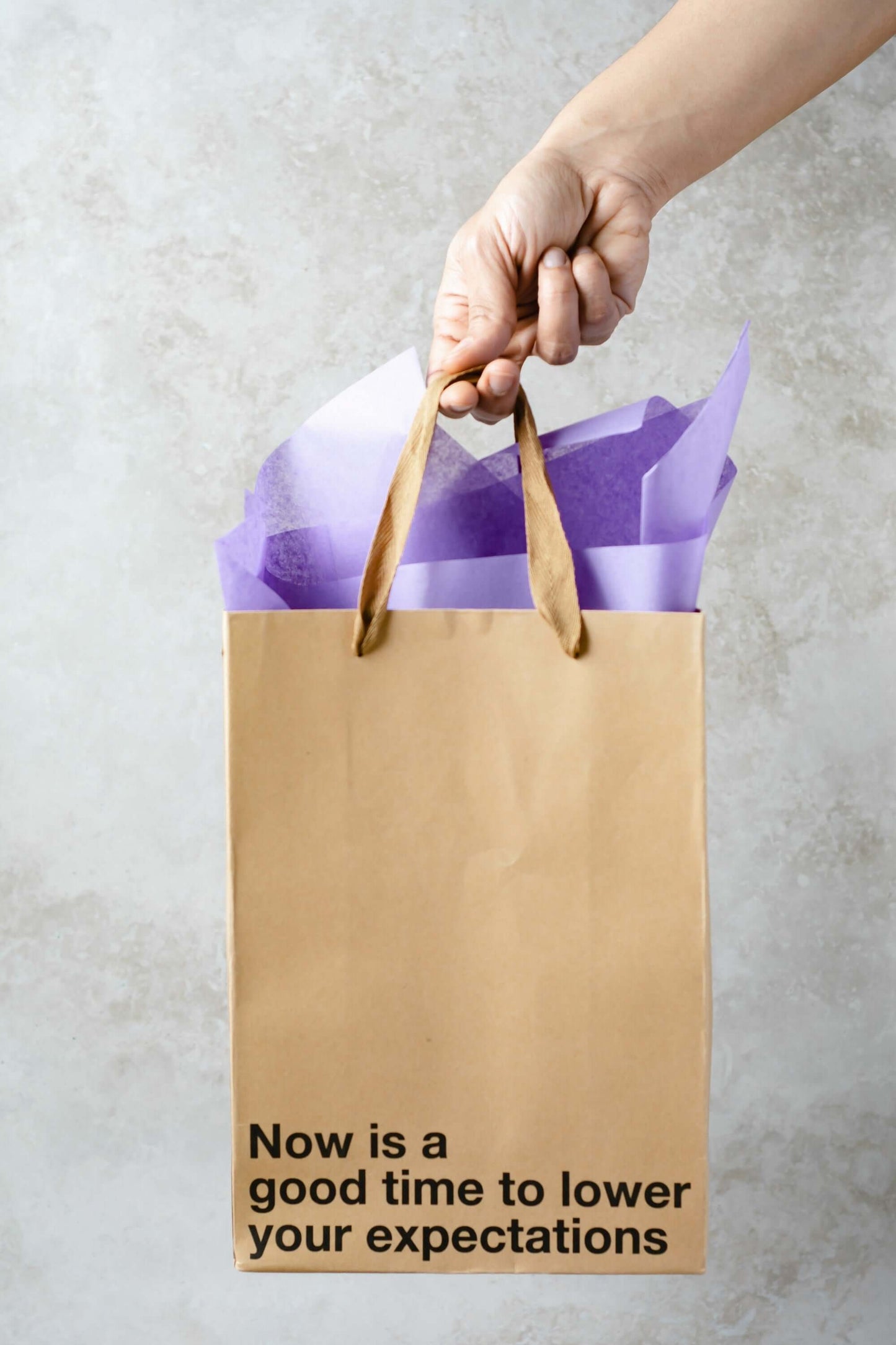 Hand holding a funny gift bag with the phrase 'Now is a good time to lower your expectations' and purple tissue paper.