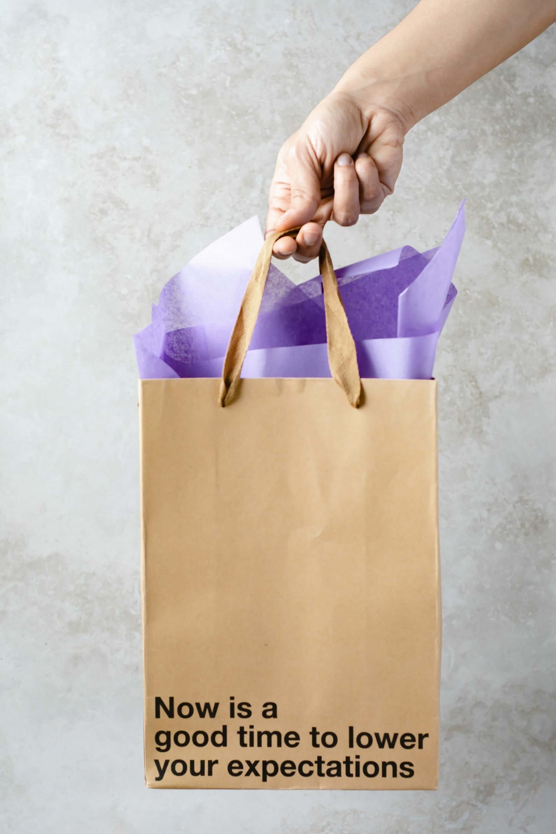 Hand holding a funny gift bag with the phrase 'Now is a good time to lower your expectations' and purple tissue paper.