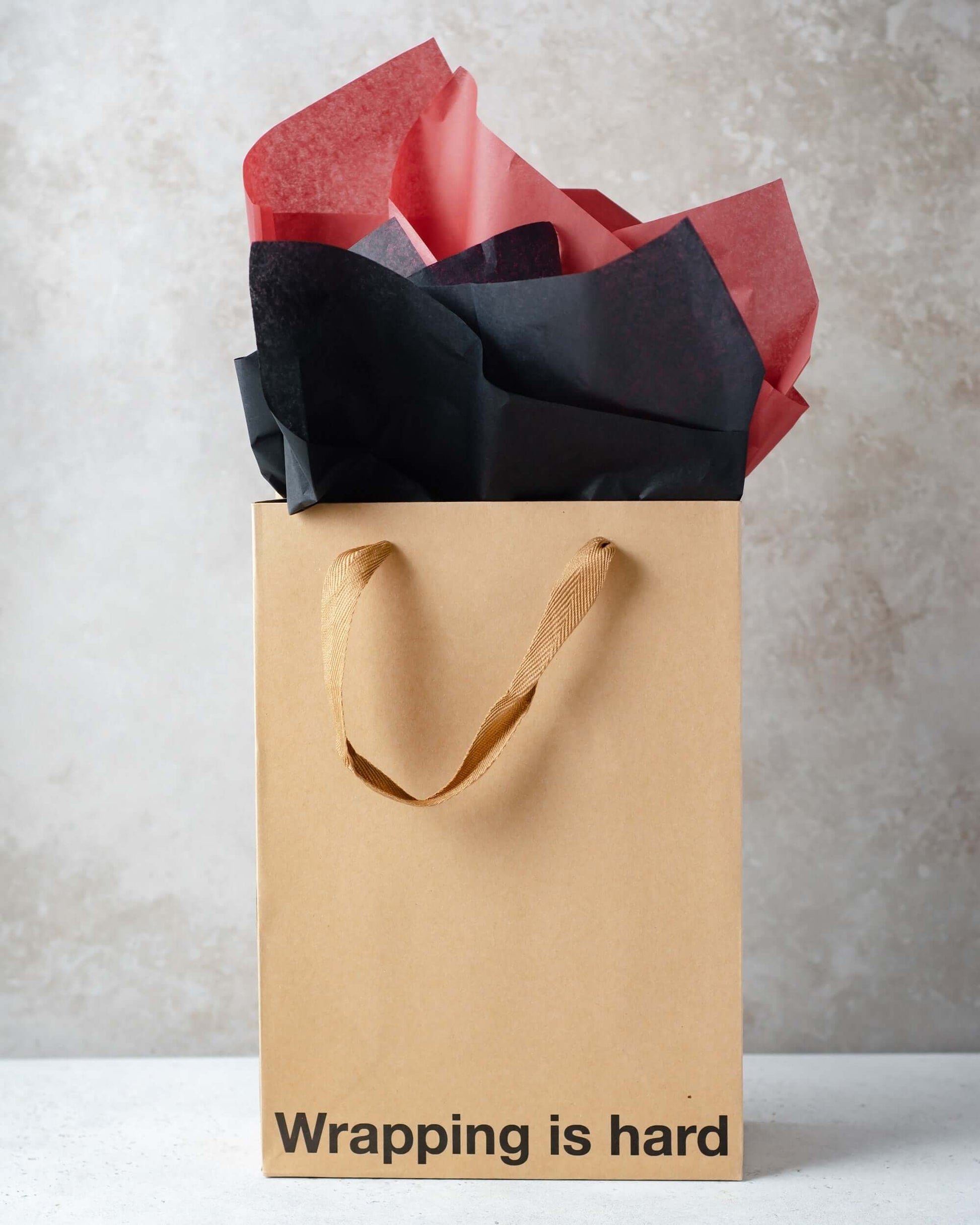 Funny gift bag with kraft material and brown ribbon handle, featuring colored tissue paper and the phrase 'Wrapping is hard'.