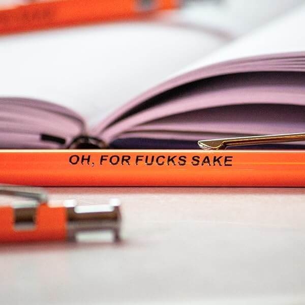 Funny pens with sarcastic text, featuring the phrase 'Oh For Fucks Sake' on an orange pen next to a notebook.