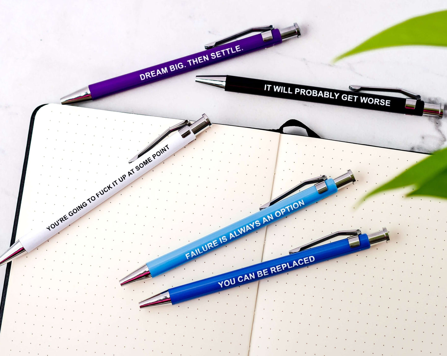 Funny pens with demotivational phrases on a notepad, including 'You can be replaced' and 'It will probably get worse'.