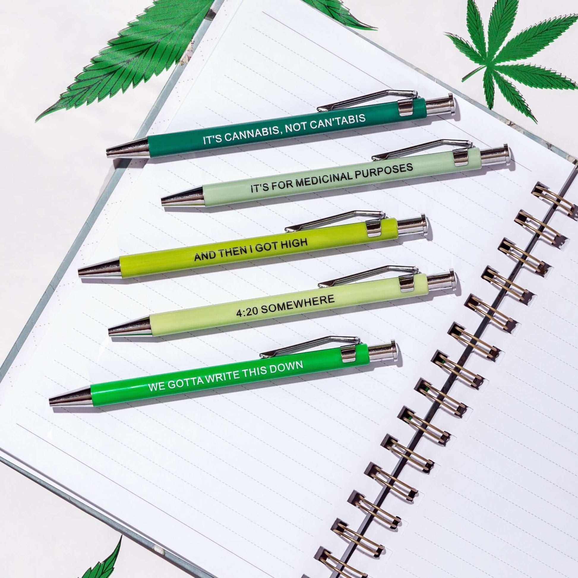 Funny pens with cannabis-related sayings on a notebook surrounded by green leaves.